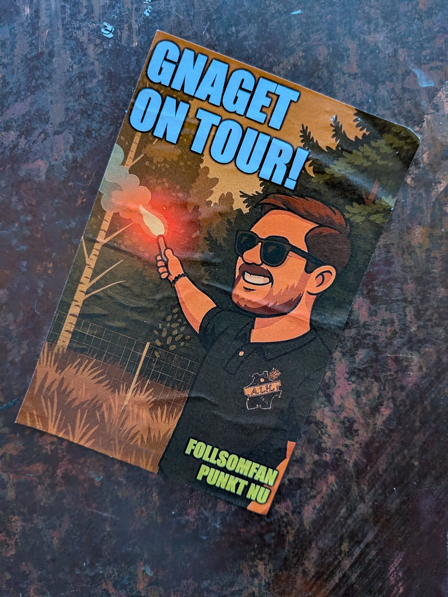 A rectangular sticker featuring a cartoon illustration of a smiling, bearded man with sunglasses, wearing a dark polo shirt with a small, intricate emblem (possibly 'A.I.H.' with a crown/lion motif). He is holding a lit red flare in his right hand, emitting white smoke. The background depicts a rustic forest scene with tall trees, including birch-like trees on the left and pine-like trees on the right, with a wire fence running through the middle ground. The color palette is dominated by earthy browns and greens, contrasted by the bright red and white of the flare. Large, light blue text with a dark outline is prominently displayed at the top, and smaller, yellow-green text on a black background is at the bottom.