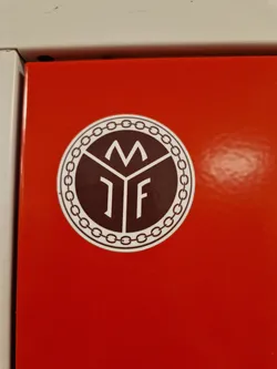 A round sticker with a dark brown center featuring the letters M and IF inside a geometric shape. It's encircled by a decorative chain-like border. The sticker is affixed to a bright red surface.
