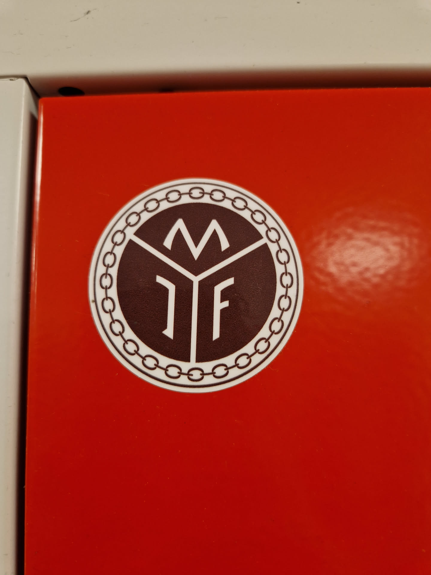 A round sticker with a dark brown center featuring the letters M and IF inside a geometric shape. It's encircled by a decorative chain-like border. The sticker is affixed to a bright red surface.