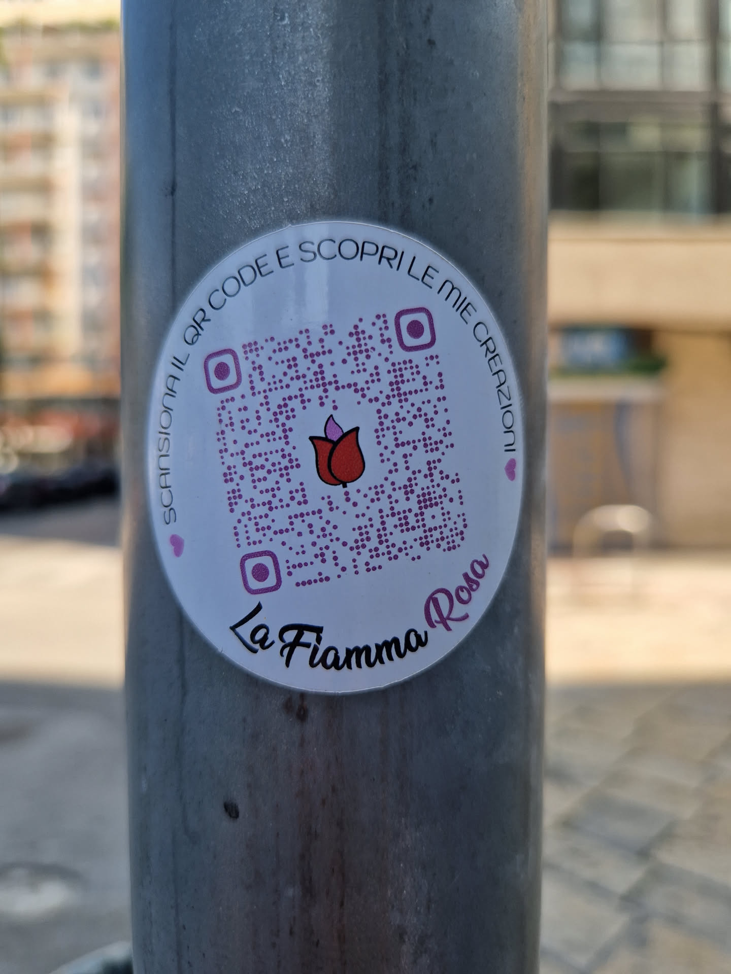 Round white sticker with a QR code in the center and the text "La Fiamma Rosa" at the bottom.  The text around the QR code says (in Italian):  "Scansiona il QR code e scopri le mie creazioni" which translates to: "Scan the QR code and discover my creations."