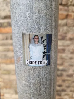 A sticker featuring a photo of a bride-to-be in a wedding dress. The photo seems to be from the past, with a slightly vintage look. The sticker is affixed to a weathered metal pole, possibly outdoors.