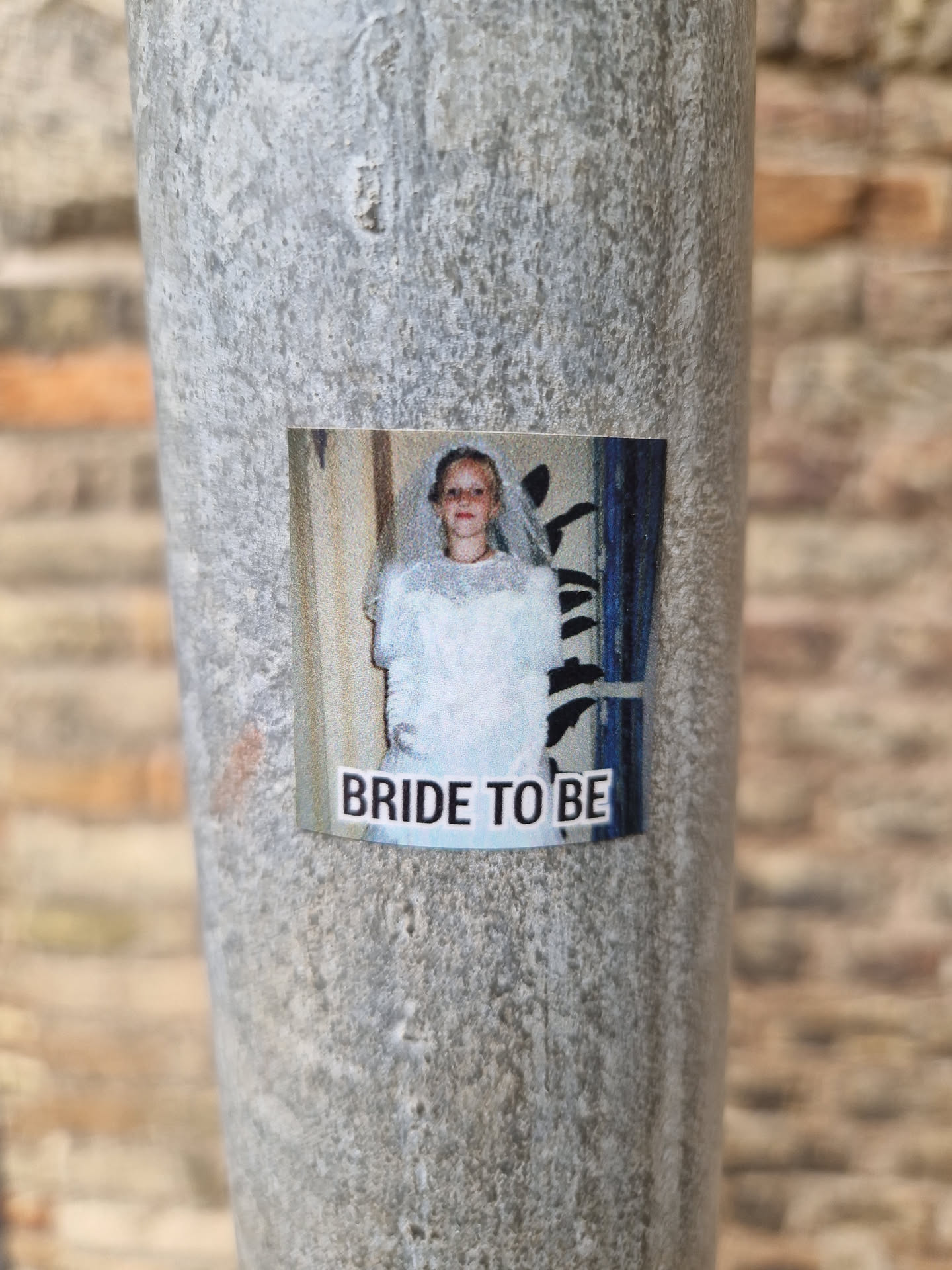 A sticker featuring a photo of a bride-to-be in a wedding dress. The photo seems to be from the past, with a slightly vintage look. The sticker is affixed to a weathered metal pole, possibly outdoors.