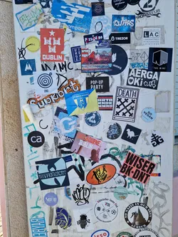 A close-up shot of a sticker featuring a red star and a castle, with the word "DUBLIN" underneath. The sticker is part of a larger collection of stickers, graffiti, and tags on a white wall.  Many of the other stickers are illegible.