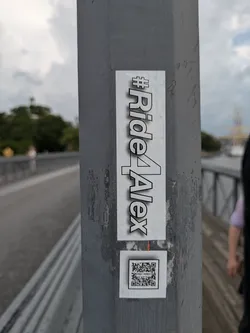 A vertically oriented rectangular sticker with a white background featuring the black text "#Ride4Alex" in a bold, sans-serif font with a subtle shadow effect. Below it, a smaller square white sticker displays a black and white QR code. Both stickers are affixed to a textured grey pole in an outdoor setting, likely a bridge or pathway, with some visible wear and slight peeling on the edges of the main sticker.