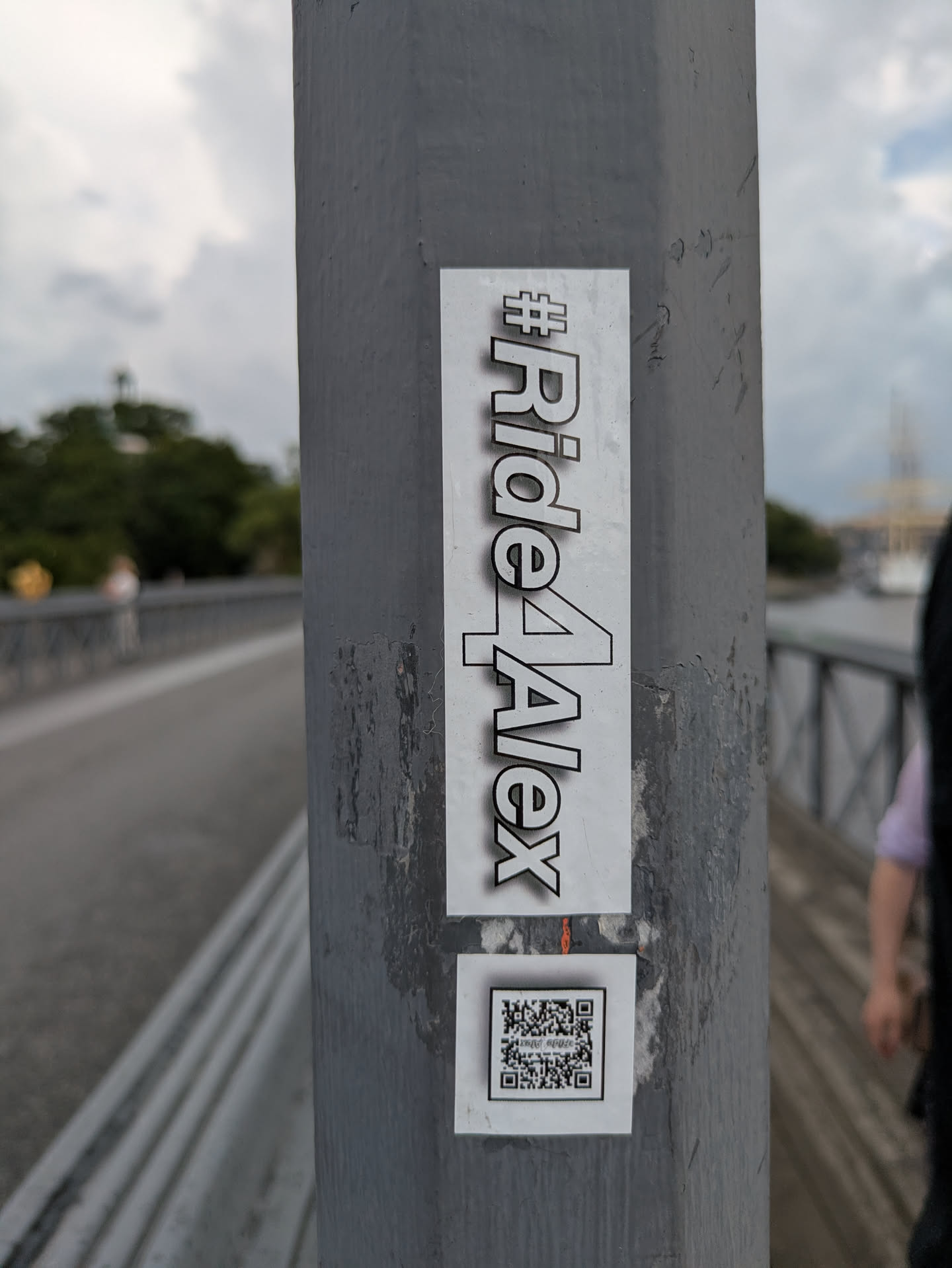 A vertically oriented rectangular sticker with a white background featuring the black text "#Ride4Alex" in a bold, sans-serif font with a subtle shadow effect. Below it, a smaller square white sticker displays a black and white QR code. Both stickers are affixed to a textured grey pole in an outdoor setting, likely a bridge or pathway, with some visible wear and slight peeling on the edges of the main sticker.