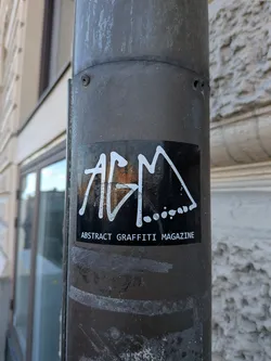 Street sticker A rectangular black sticker attached to a cylindrical weathered metal pole. The center features a bold, white graffiti handstyle tag spelling out the letters 'AGM', with decorative stylized lines and a row of trailing dots connected to the 'M'. Directly beneath the tag, clear white uppercase sans-serif text reads 'ABSTRACT GRAFFITI MAGAZINE'.