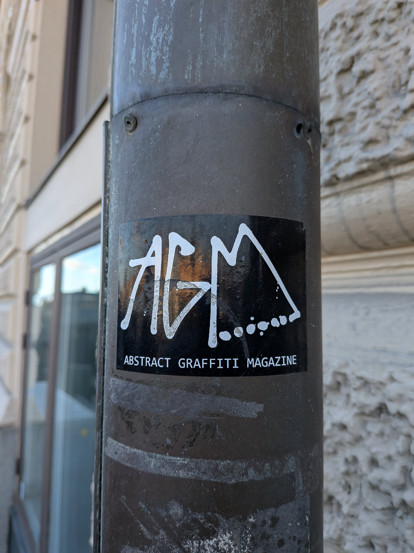 A rectangular black sticker attached to a cylindrical weathered metal pole. The center features a bold, white graffiti handstyle tag spelling out the letters 'AGM', with decorative stylized lines and a row of trailing dots connected to the 'M'. Directly beneath the tag, clear white uppercase sans-serif text reads 'ABSTRACT GRAFFITI MAGAZINE'.