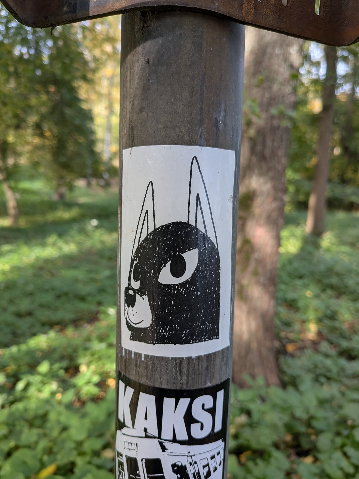 The sticker is predominantly white with a black illustration. It depicts the head of a dog or a similar canine creature. The face is filled with a textured black pattern, resembling a rough sketch or a stippled effect. Two large, oval eyes with black pupils are prominent. Above the head, two long, pointed ears extend upwards. A small, simple nose and a subtle smile are also visible. The sticker has a clean white border.