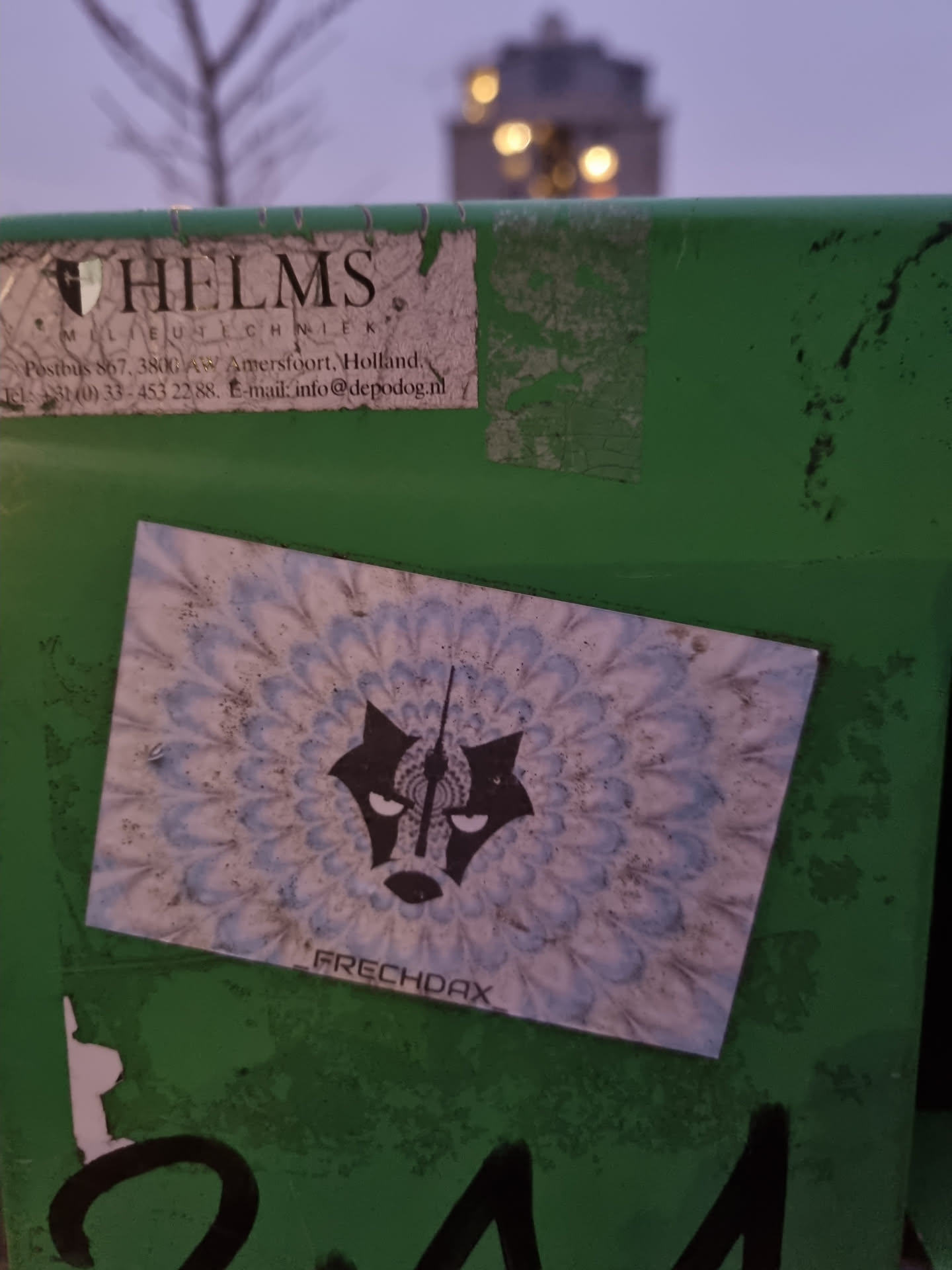 A sticker with a black wolf head in the center of a mandala-like pattern. The word "FRECHDAX" is printed under the design. The sticker is affixed to a green surface.