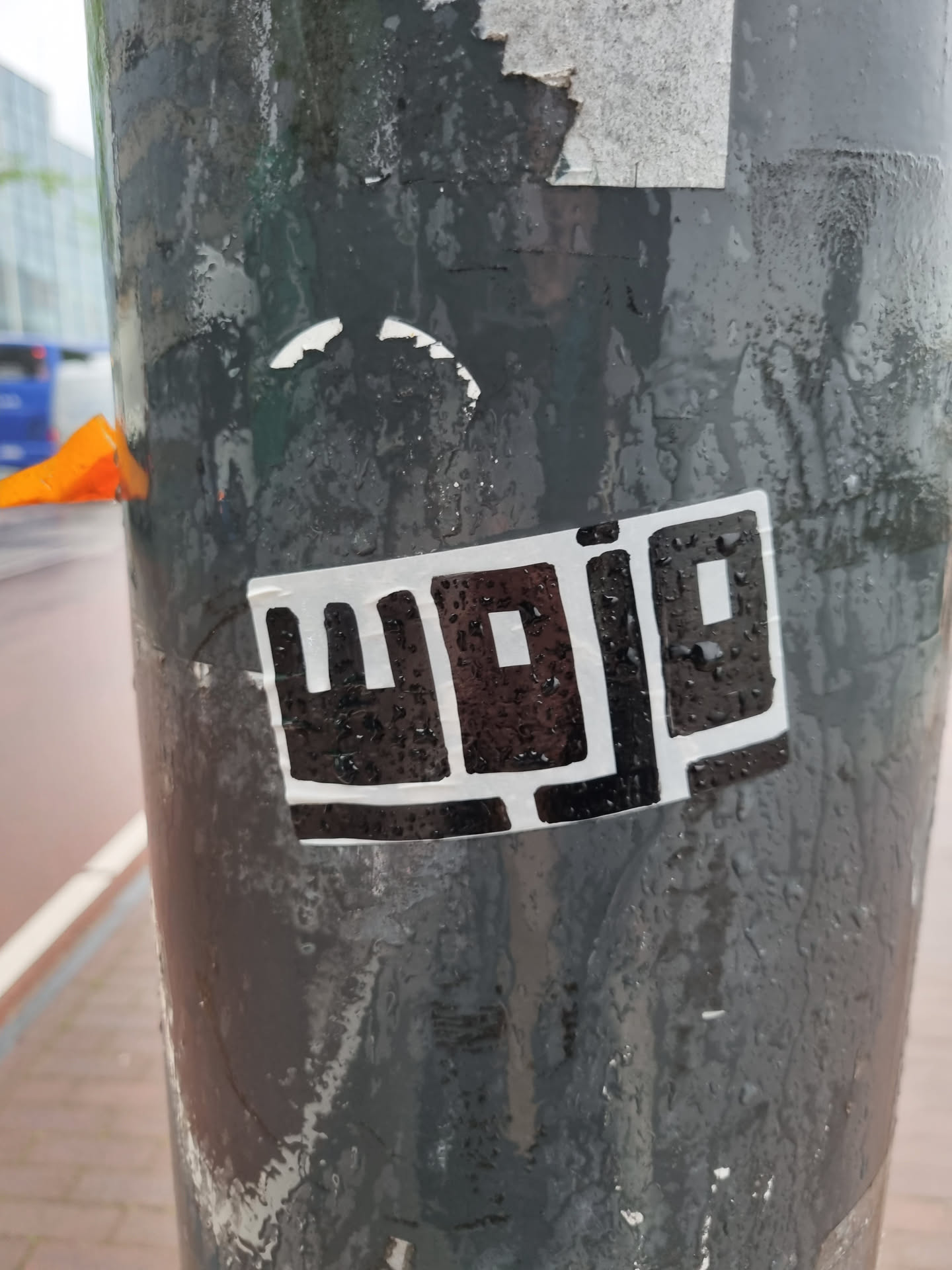 A black and white sticker with an abstract design. The sticker is affixed to a weathered pole, with rain droplets visible on its surface. The main visual element appears to be a stylized logo or wordmark, possibly an abbreviation, though not fully legible.