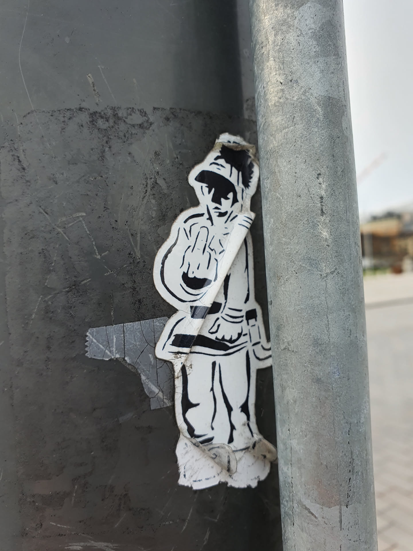 A monochrome sticker featuring a stylized figure in uniform, possibly military or historical, making a rude gesture. The sticker is affixed to a metallic pole, showing signs of wear and tear.