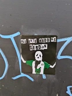 This sticker depicts a cartoonish skeleton figure wearing a green and white football jersey, with arms raised in celebration. Text above the figure reads 'BLI INTE RÄDD NU GÅRDAN'. The sticker is placed on a grey wall with light blue graffiti nearby.