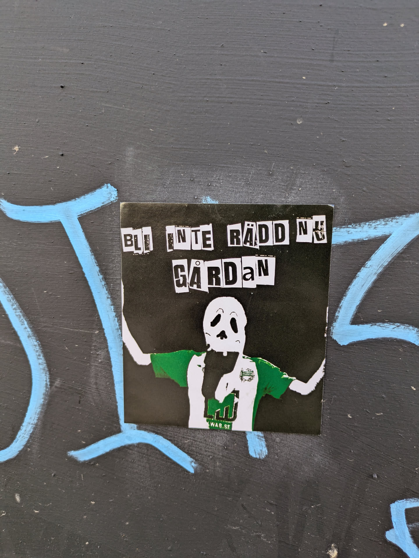 This sticker depicts a cartoonish skeleton figure wearing a green and white football jersey, with arms raised in celebration. Text above the figure reads 'BLI INTE RÄDD NU GÅRDAN'. The sticker is placed on a grey wall with light blue graffiti nearby.