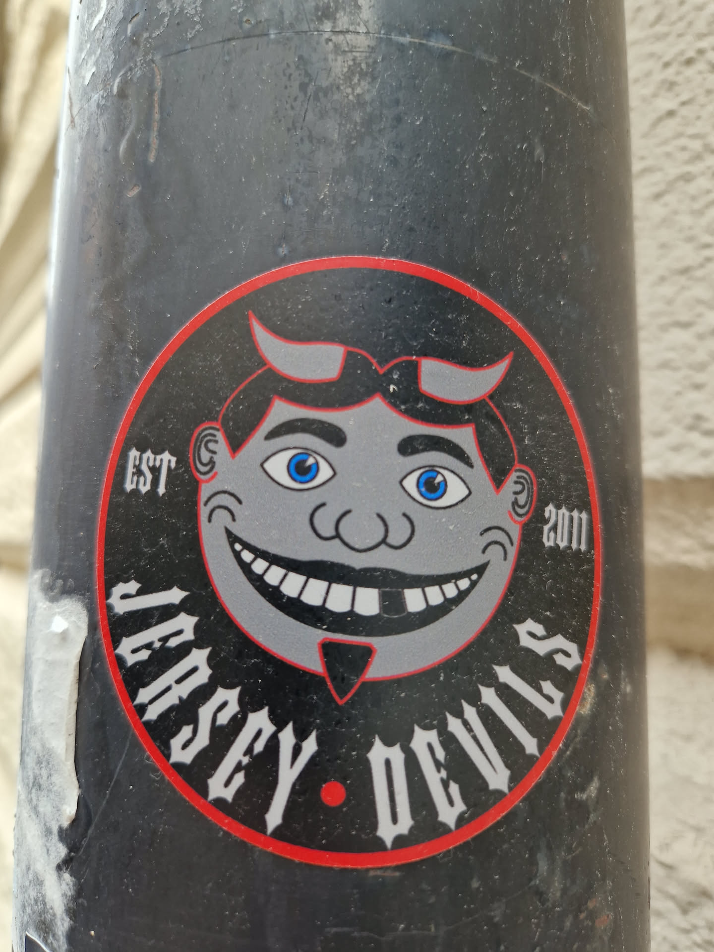 A round sticker featuring a cartoon devilish face with horns and a mischievous grin. The text 'Jersey Devils' is written in a stylized font around the character. The years 'EST 2011' are subtly included in the design. The sticker is adhered to a dark grey cylindrical surface. 