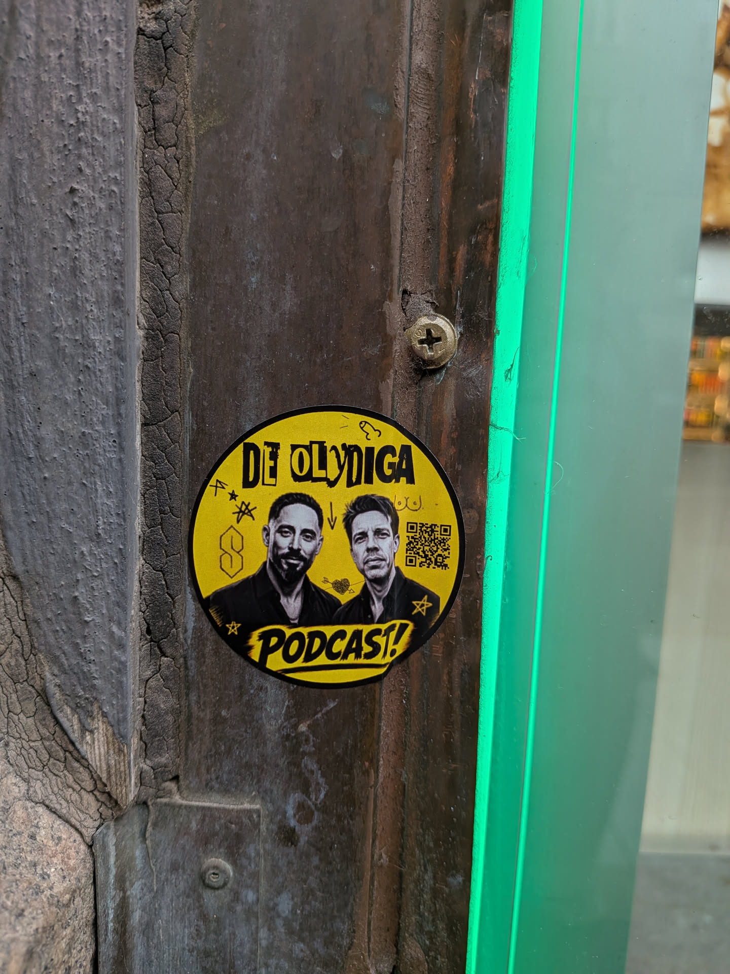 A circular yellow sticker affixed to a dark, weathered wooden or metal surface. The sticker features black and white photographic portraits of two men in the center. Above them, the text 'DE OLYDIGA' is printed in a bold, stylized black font. Below them, the word 'PODCAST!' appears in a similar font within a yellow banner. The background is filled with various small black doodles, including a 'Cool S' symbol, stars, an arrow, a heart with an arrow, and crude drawings of male and female anatomy. A QR code is visible on the right side of the sticker.