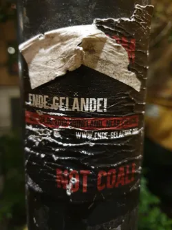 A worn sticker on a pole with the text Ende Gelände! and No Coal!. The sticker shows some damage and is partially peeled.  The website www.ende-gelände.org is visible.