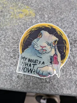 A sticker featuring a rabbit with a skeptical expression. The rabbit is depicted in a circular frame with a dark background. The sticker has a holographic effect. The text 'My what's a what now?' is written underneath the rabbit.