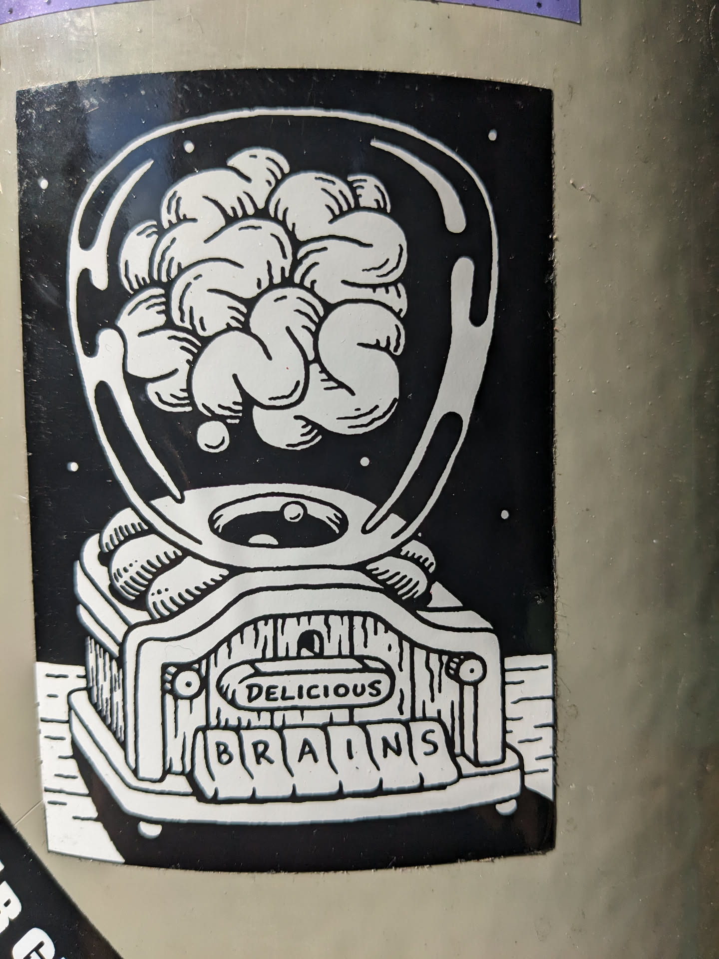A black and white sticker depicting a brain inside a glass dome on top of a machine with the words "Delicious Brains" written on it.