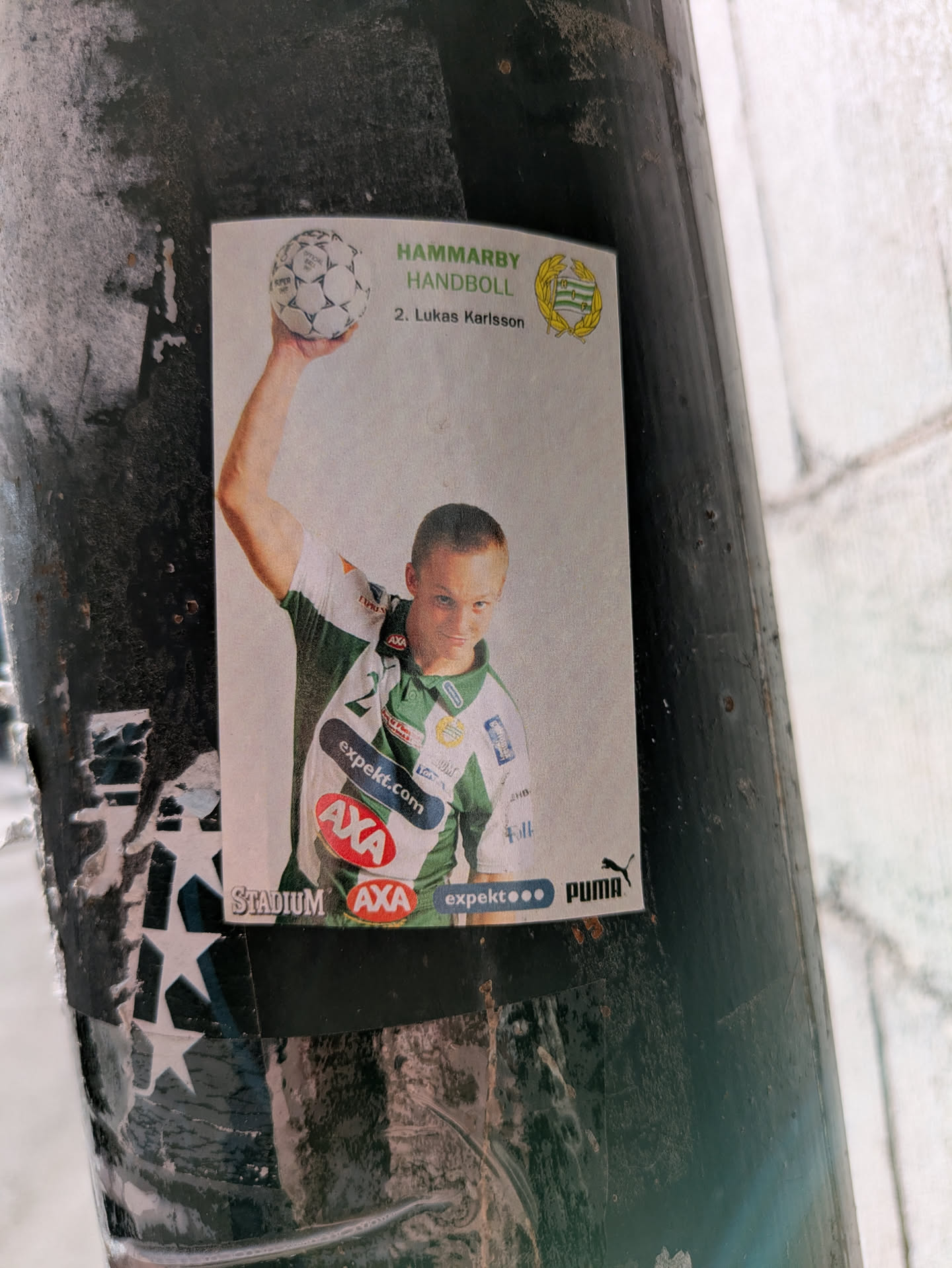 The sticker features a picture of Lukas Karlsson, a handball player from Hammarby Handboll, a Swedish handball team. He is holding a handball in his right hand and wearing the team's jersey. The sticker contains logos of sponsors such as Stadium, AXA, expekt.com and Puma.