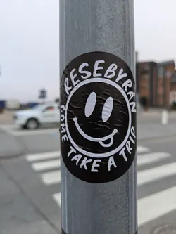 A black and white sticker with a smiley face and the words "ReseByran" and "Come take a trip" around it.  The sticker is affixed to a metal pole.