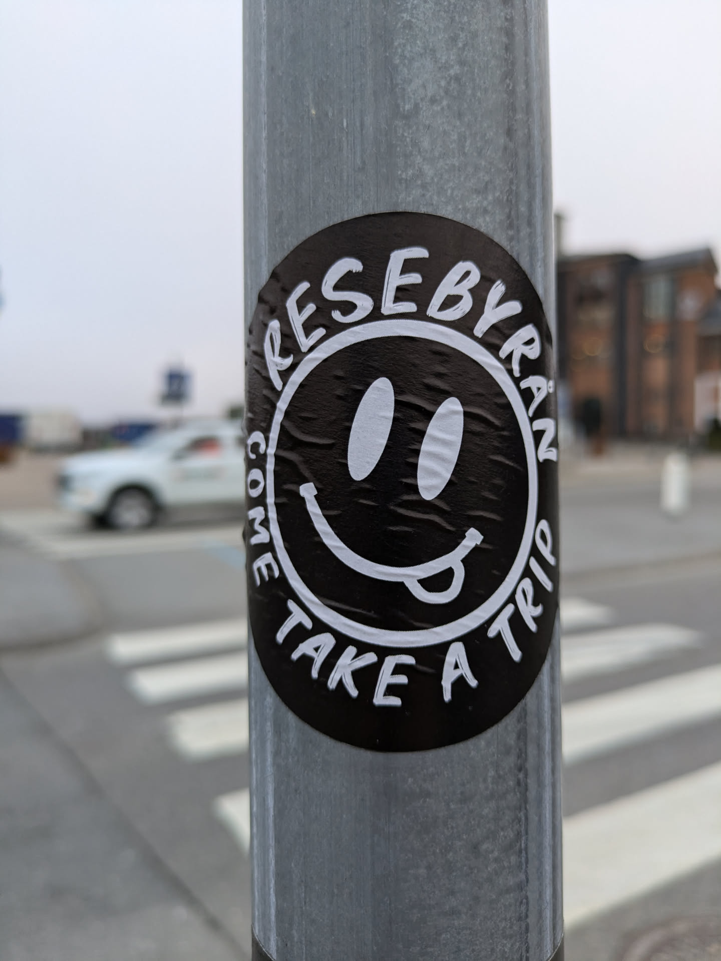 A black and white sticker with a smiley face and the words "ReseByran" and "Come take a trip" around it.  The sticker is affixed to a metal pole.
