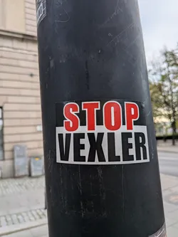 Street sticker A rectangular sticker stuck onto a black pole. The top half has a black background displaying the word 'STOP' in tall, bold red block letters outlined in thin white. The bottom half features a light gray/white background with the word 'VEXLER' printed in tall, condensed black capital letters.