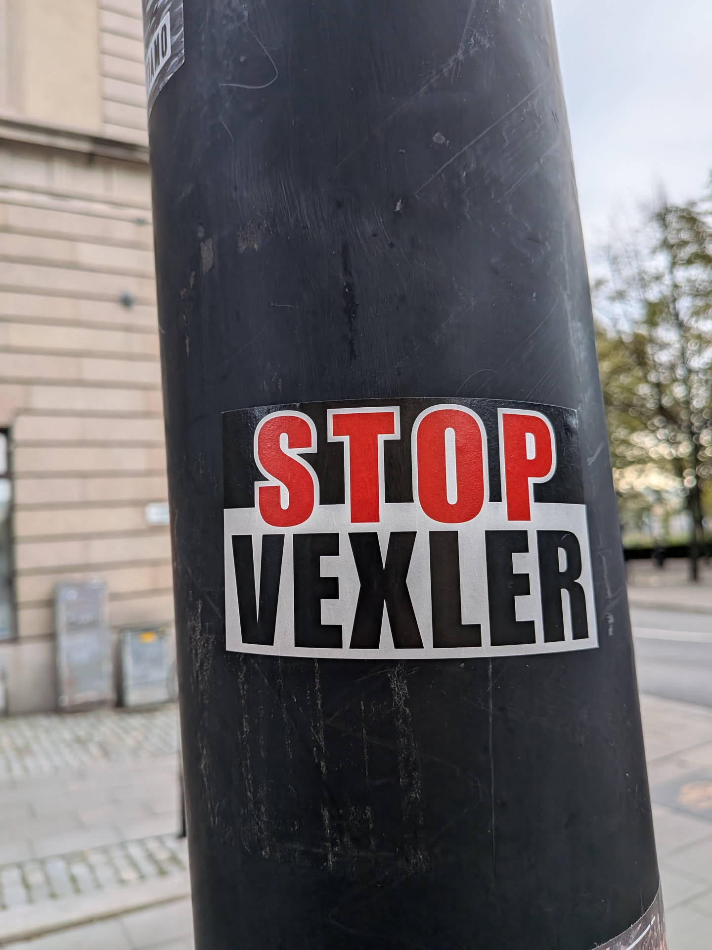 A rectangular sticker stuck onto a black pole. The top half has a black background displaying the word 'STOP' in tall, bold red block letters outlined in thin white. The bottom half features a light gray/white background with the word 'VEXLER' printed in tall, condensed black capital letters.