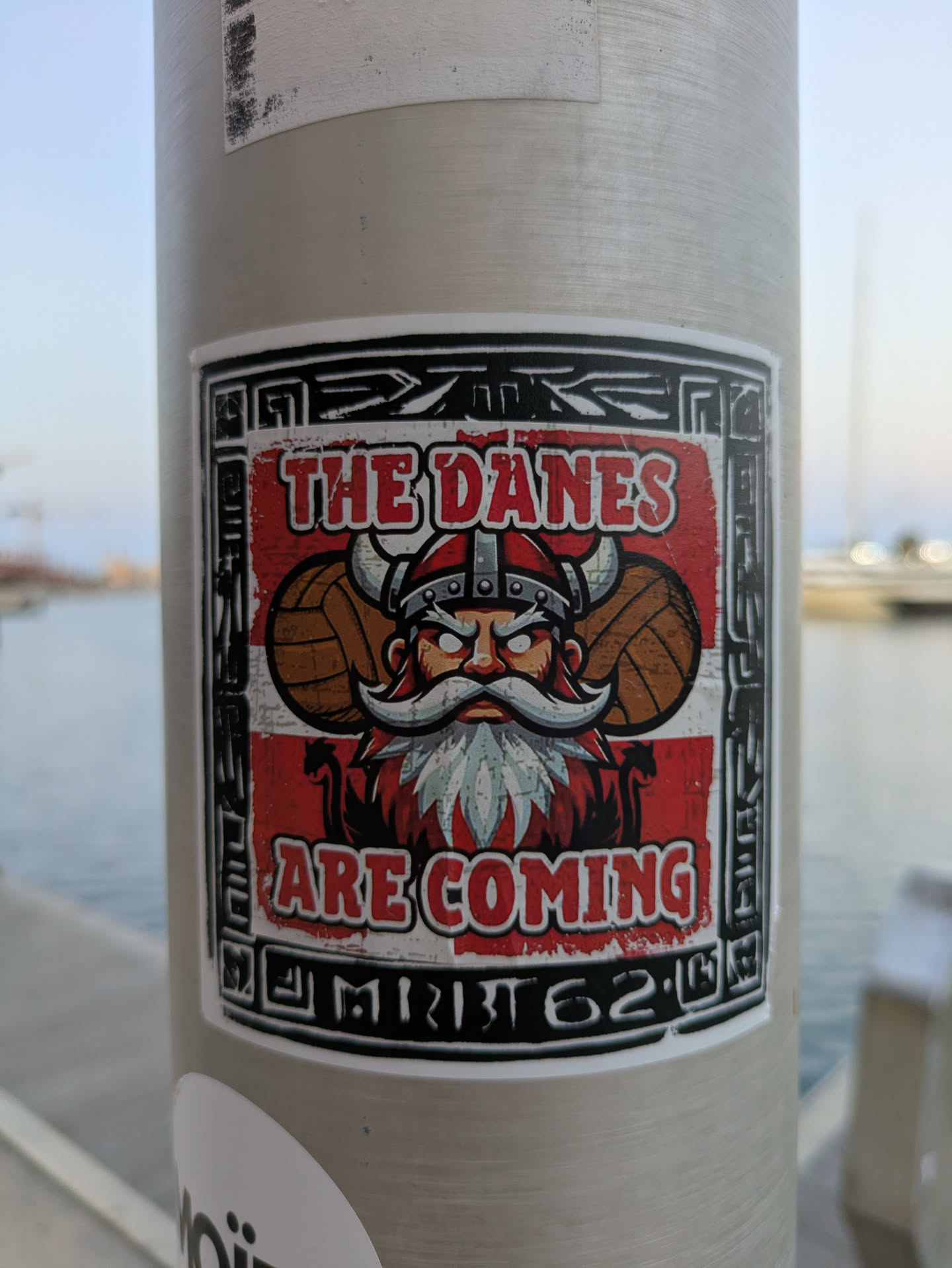 The sticker is square with a white border and the image features a viking with a red and white background reminiscent of the Danish flag. The viking has a horned helmet with what appears to be footballs on either side, a large white mustache and beard, and a determined expression. Below the viking is the text 'ARE COMING' in bold, red letters, with the word 'M1B162' under it.