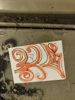 A square sticker with an abstract design in orange and white. The design includes swirls, hearts, and geometric shapes. It's attached to a dirty, metallic surface.