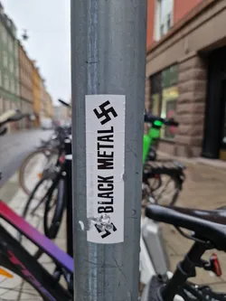 A sticker with the text "Black Metal" and a reversed swastika symbol attached to a metal pole. The background is a city street with parked bicycles.