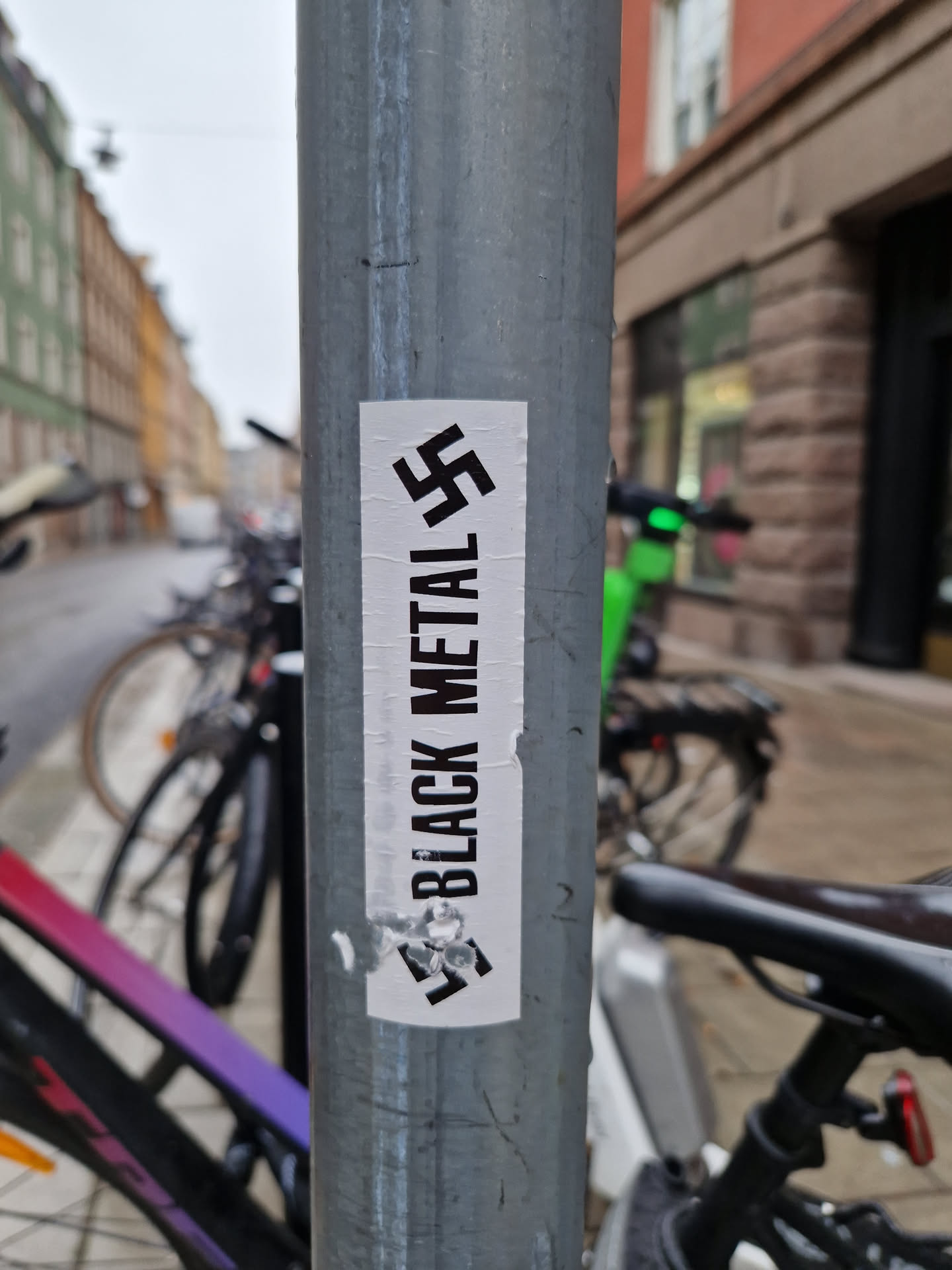 A sticker with the text "Black Metal" and a reversed swastika symbol attached to a metal pole. The background is a city street with parked bicycles.