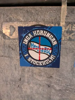 A square sticker predominantly in shades of blue, white, and black, featuring a round emblem in the center. The circular border contains the Swedish text 'INGA HORUNGAR' on top and 'I STOCKHOLM' on the bottom in stylized block lettering with black borders. In the middle of the emblem, the Hertha BSC flag/text is positioned in the center of a red crosshair target reticle against a blue and white patterned background.