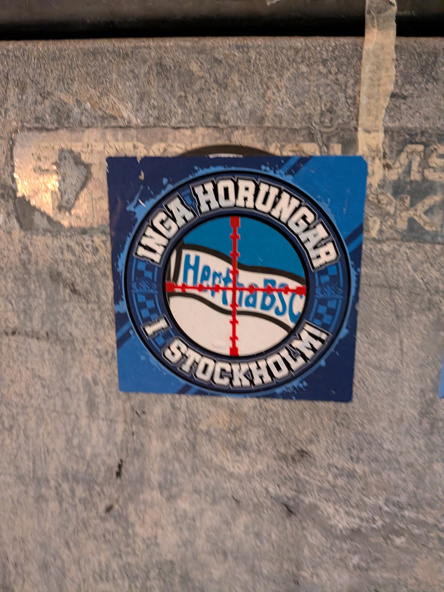 A square sticker predominantly in shades of blue, white, and black, featuring a round emblem in the center. The circular border contains the Swedish text 'INGA HORUNGAR' on top and 'I STOCKHOLM' on the bottom in stylized block lettering with black borders. In the middle of the emblem, the Hertha BSC flag/text is positioned in the center of a red crosshair target reticle against a blue and white patterned background.