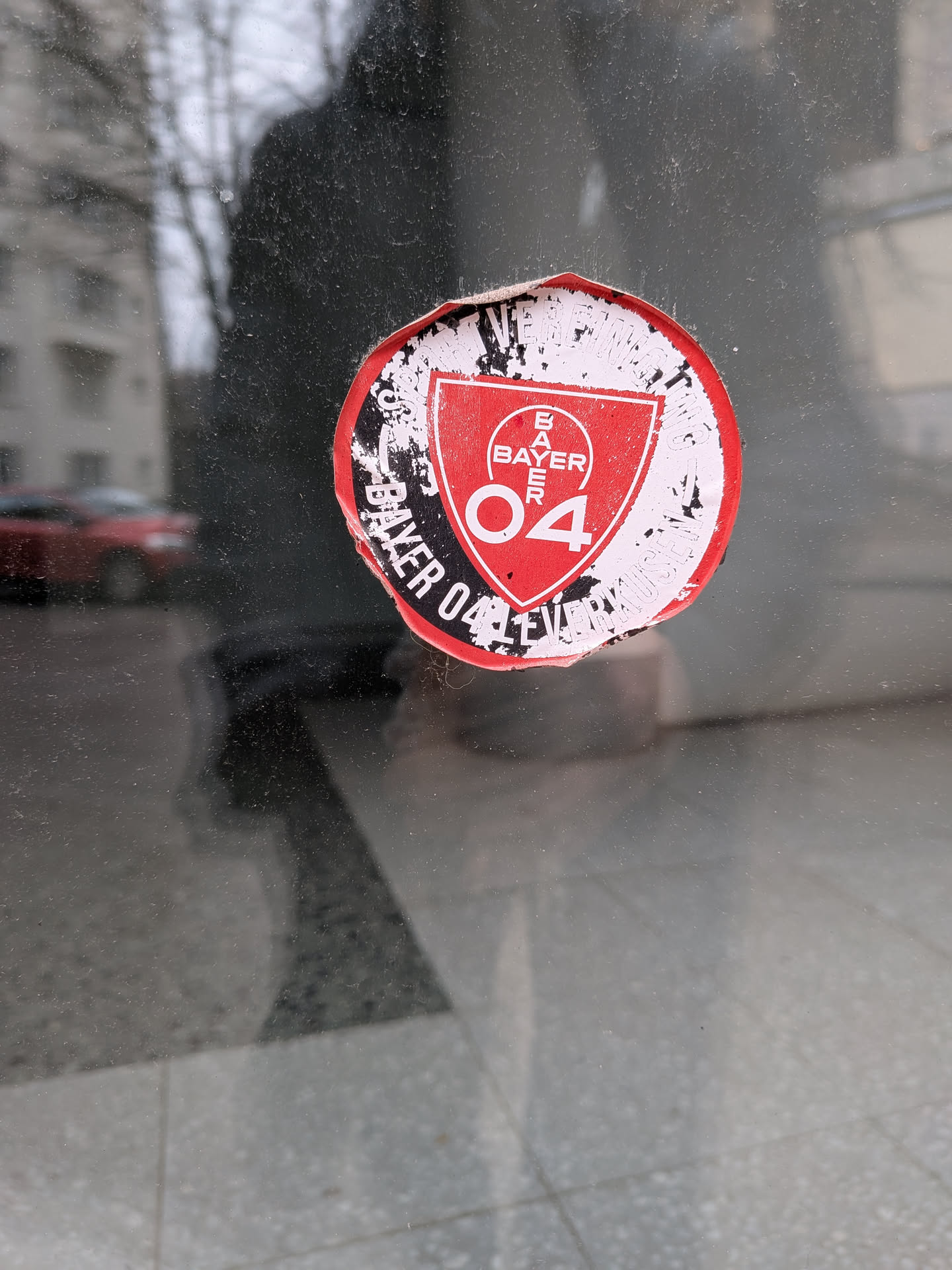 A circular sticker featuring the emblem of Bayer 04 Leverkusen. The central design is a red shield with a white outline, containing the word 'BAYER' stacked vertically in white, topped by the Bayer cross logo, and the number '04' in white below it. The shield is set against a black background, which is then surrounded by a white ring. Around the outer edge of the white ring, in black text, are the words 'BAYER 04 LEVERKUSEN VEREIN FÜR LEIBESÜBUNGEN'. The sticker shows significant signs of wear and tear, with frayed edges, peeling, and dirt accumulation, particularly around the black and white outer ring, giving it a distressed and aged appearance.