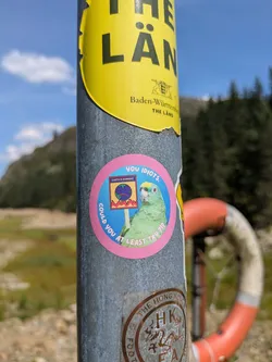 A circular sticker affixed to a metal pole features a vibrant green parrot with a yellow head, holding a sign. The sign depicts a sad blue globe with red flames underneath, bearing the text 'EARTH IS BURNING'. Surrounding the central image, in a pink border, the text 'you IDIOTS.' is at the top, and 'COULD YOU AT LEAST TRY ?!?' is curved along the bottom. The background shows a natural outdoor setting with a mountain, trees, and a lifebuoy.