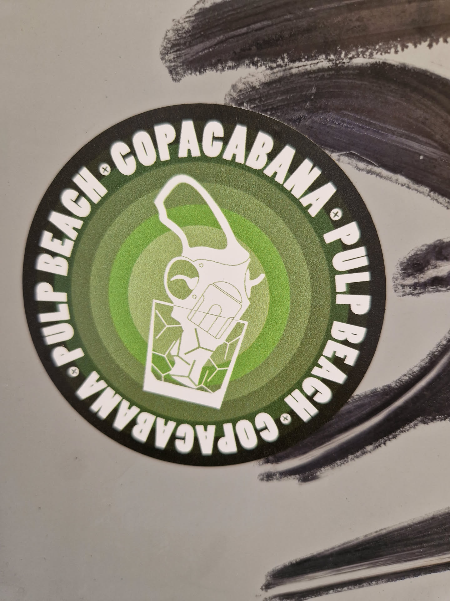 Round sticker with a green and white design. It features a drawing of a cocktail shaker pouring a drink into a glass, encircled by the text "Pulp Beach Copacabana" repeated twice in a circular fashion.