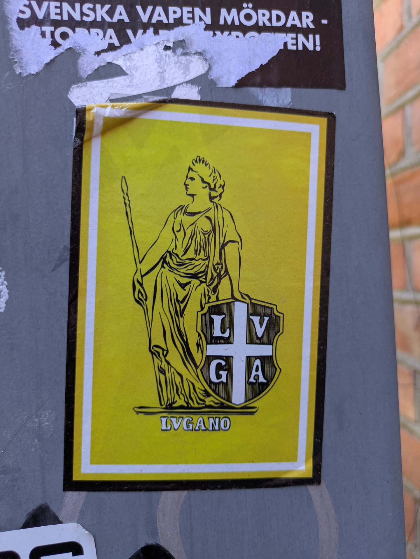 This is a sticker on a wall. It features a yellow background, framed by a white border. The primary image is a drawing of a woman in a classical style, wearing a crown and holding a staff. Next to her is a shield with the letters "LVGA" on it. Below, the word "LVGANO" is printed.