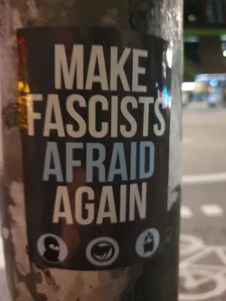A sticker with the text "Make fascists afraid again". It also features three symbols: a balaclava, a symbol resembling a fist, and a burning trash can. The sticker is affixed to a pole.