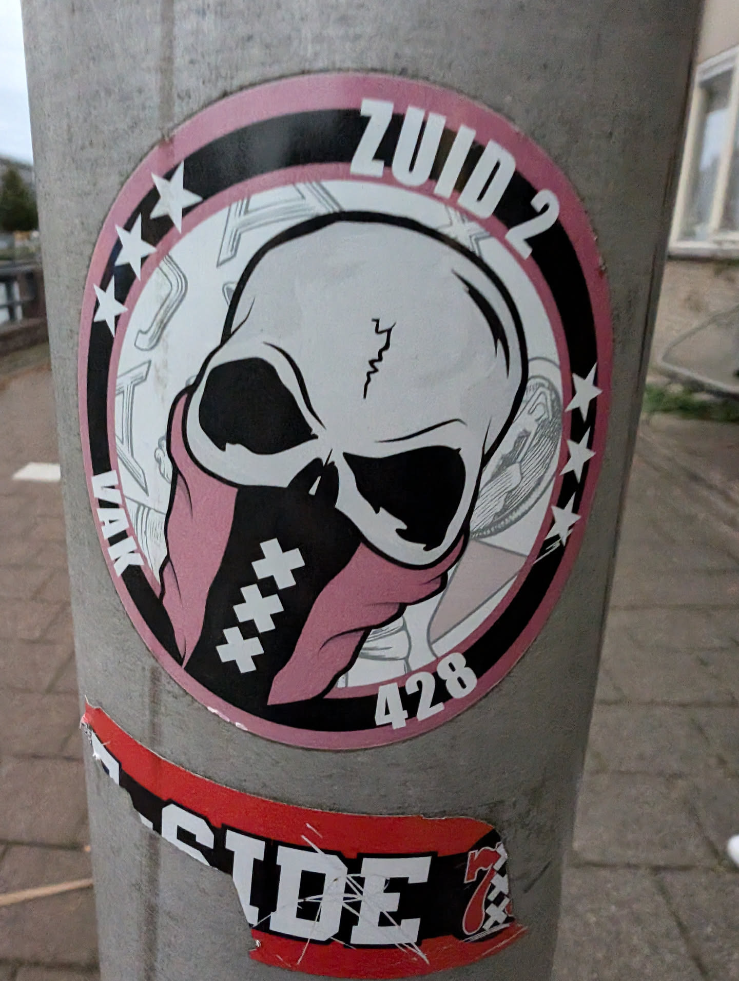 Round sticker featuring a skull wearing a pink bandana with crosses. The text ZUID 2 and 428 are visible.  The sticker has a black and pink color scheme. 