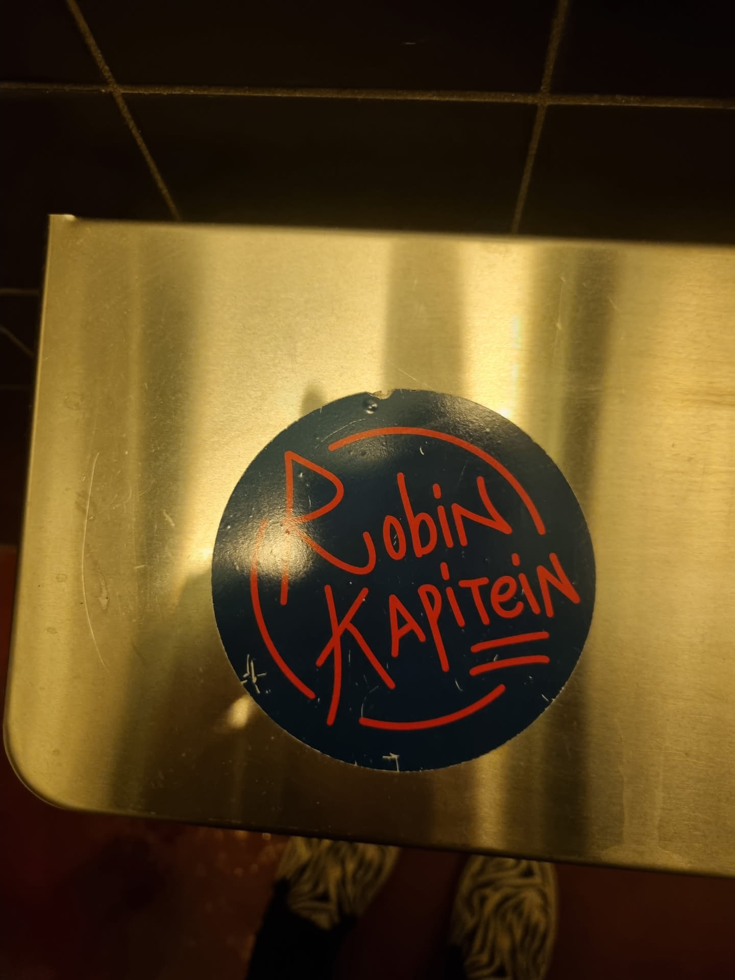 Round sticker on a metallic surface. It has a dark blue background with red lettering that says "Robin Kapitein". There's also a small graphic element resembling a ship's arrow or a stylized bird.