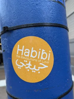 A round yellow sticker on a blue pole. The sticker says Habibi in English and Arabic script, with the tagline "The Revolutions of Love".