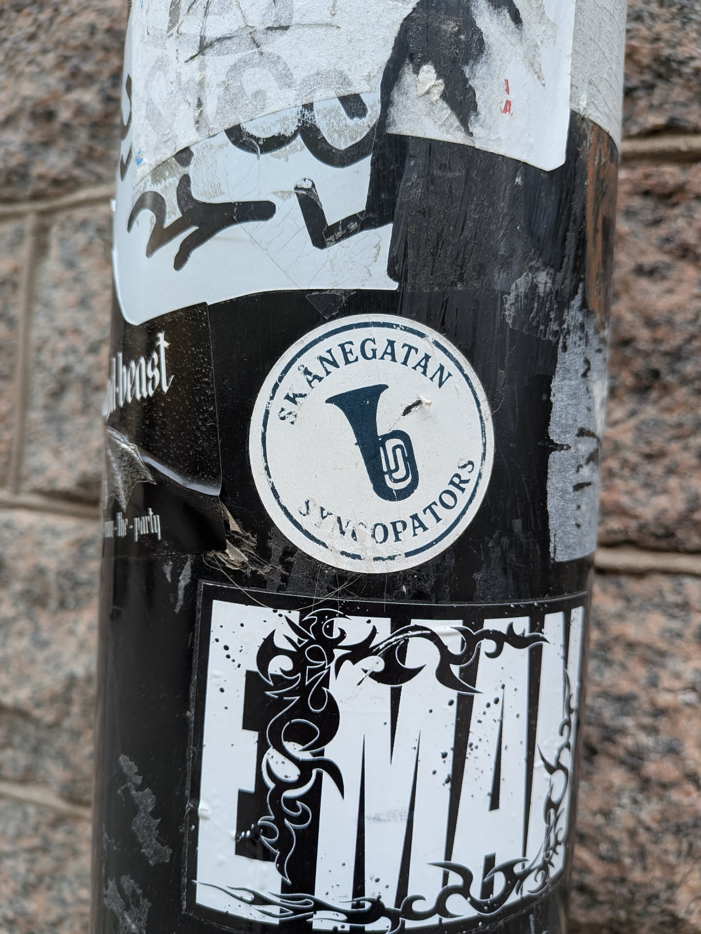 A circular sticker featuring a stylized dark blue brass instrument, resembling a tuba or baritone horn, centered on a white background. The text 'SKÅNEGATAN' is curved along the top edge of the circle, and 'SYNCOPATORS' is curved along the bottom edge, both in dark blue capital letters. A thin dark blue line forms the outer border of the circular design. The sticker shows signs of wear and tear, with some dirt and slight discoloration, indicating it has been exposed to the elements.