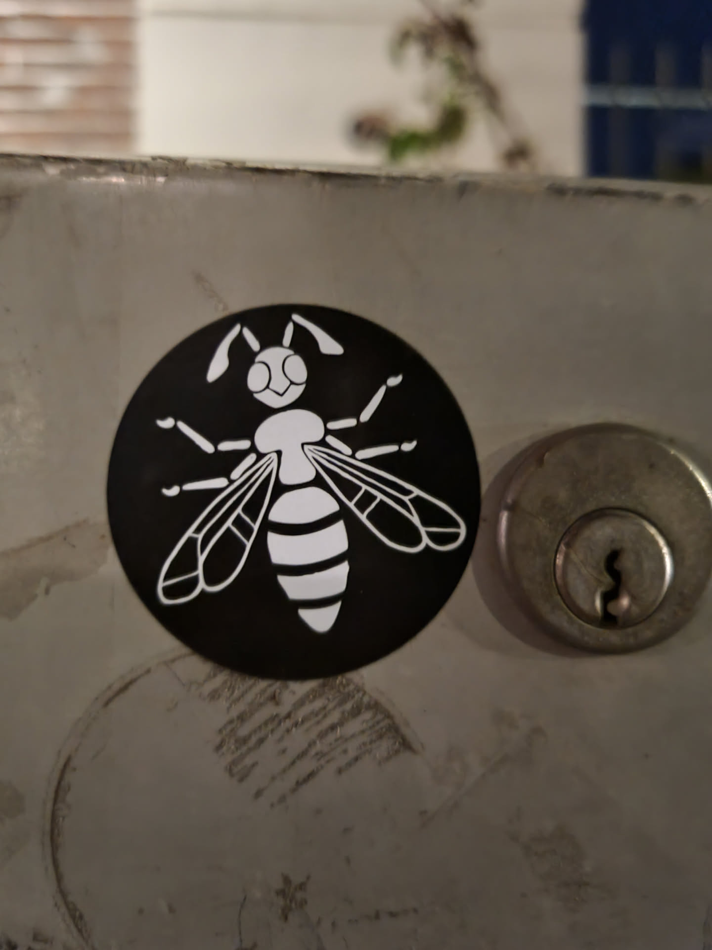 A round black and white sticker with an illustration of a bee on a dark background. The bee is depicted in a simple, almost cartoonish style, and is primarily white with some black details. The sticker is affixed to a metallic surface.