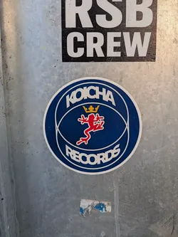 Street sticker A round sticker featuring a deep blue background with a white outer border. In the center is a stylized red frog outlined in white, topped by a small yellow crown, framed by intersecting thin white curved lines forming an oval globe-like pattern. Arced across the top in bold white capital letters is the word 'KOICHA', and arced across the bottom in identical font is 'RECORDS'.