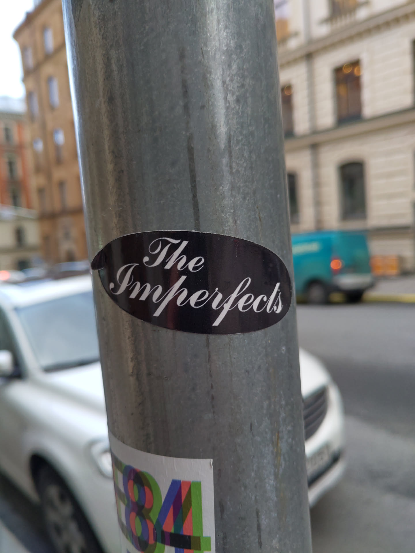 A black oval sticker with the text 'The Imperfects' in a stylized script font. It's affixed to a metal pole outdoors.