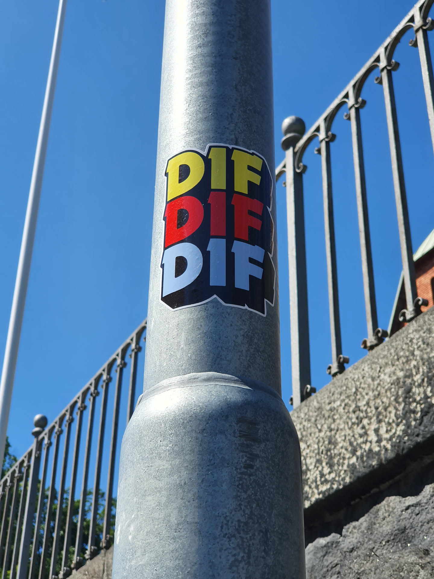 A sticker featuring the three-times repeated acronym DIF in yellow, red and light blue, on a grey metal post. The sticker has a slightly 3D effect and is applied to a metal post outdoors.