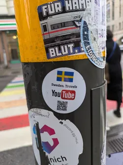 A round sticker featuring the Swedish flag and the YouTube logo.  The text "Sweden. Travels" is above the logo, and a QR code is below it. The sticker is attached to a pole covered in other stickers.