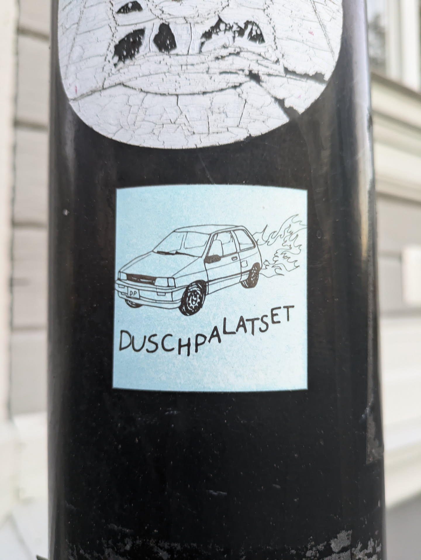 A light blue square sticker with a black line drawing of a car and flames coming out of the exhaust. The text "Duschpalatset" is written underneath the drawing.