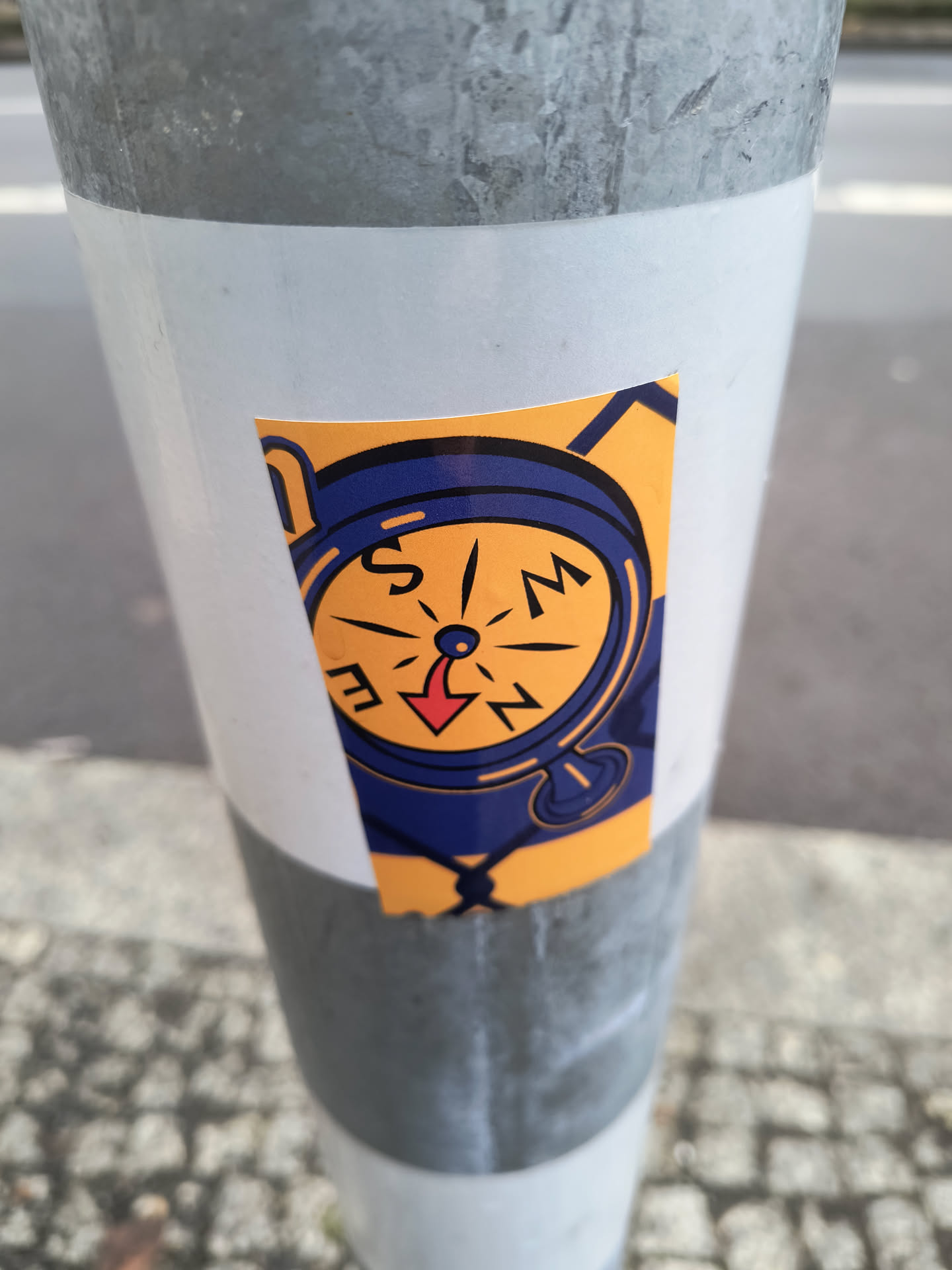 A yellow and blue illustrated compass sticker with a red arrow pointing south. It is applied to a grey pole.