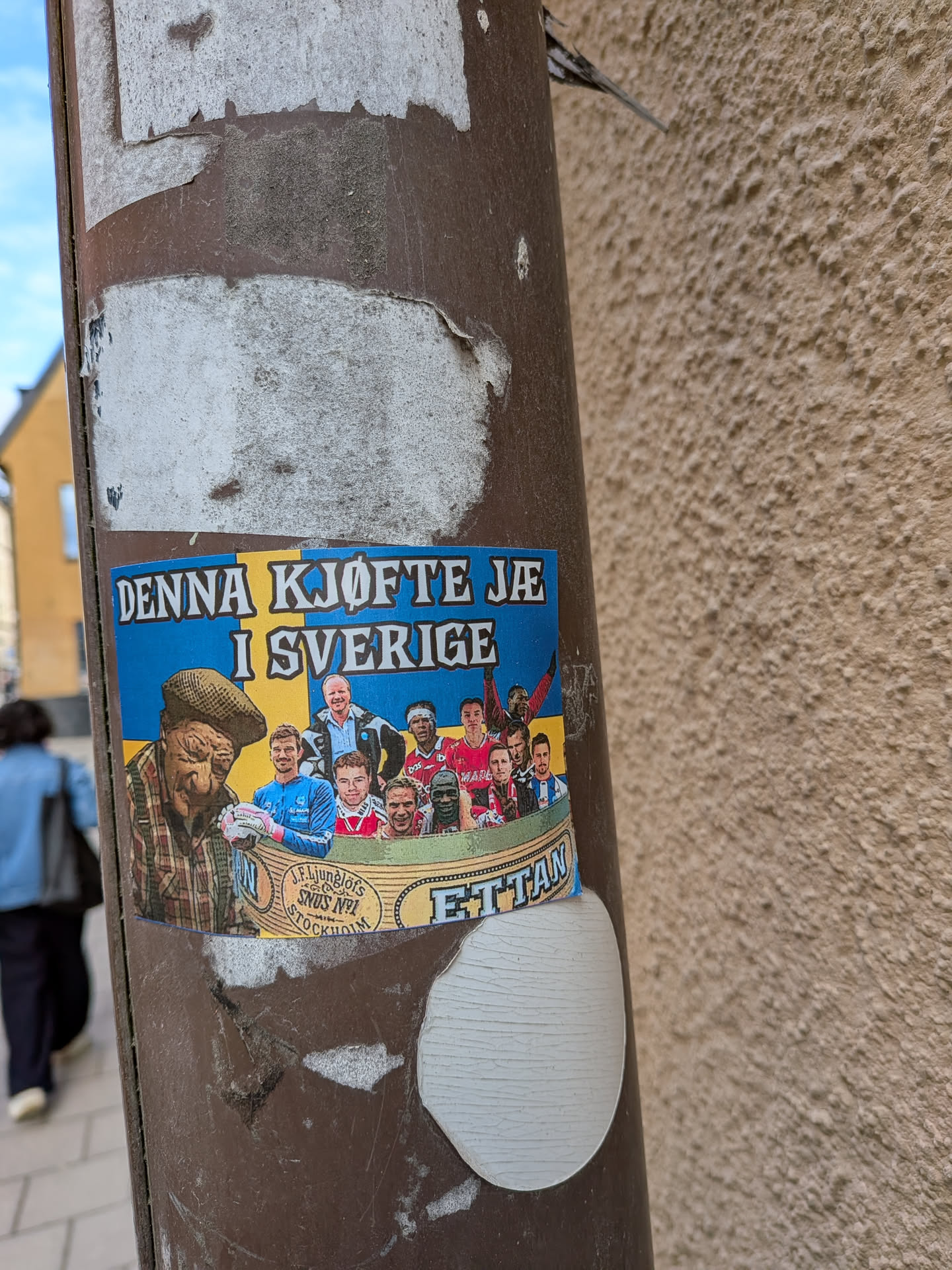The sticker is rectangular with a background mimicking the blue and yellow of the Swedish flag. On the left, an older man with a weathered face, wearing a plaid shirt, suspenders, and a flat cap, holds what appears to be a snus can. To his right, a diverse group of male football players, some in jerseys (red, blue, white), are depicted in a semi-circle, as if in a boat or a celebratory huddle. One player in a blue jersey holds a football. A curved banner at the bottom features text related to snus. The overall style is illustrative and somewhat cartoonish, combining elements of traditional Swedish identity with modern sports.